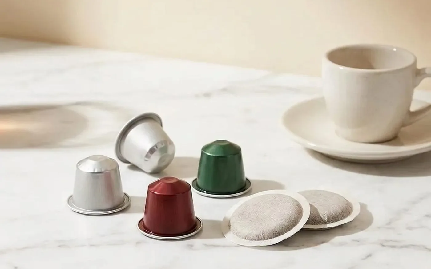 Aluminium coffee capsules and ESE paper pods arranged on white marble countertop with espresso cup