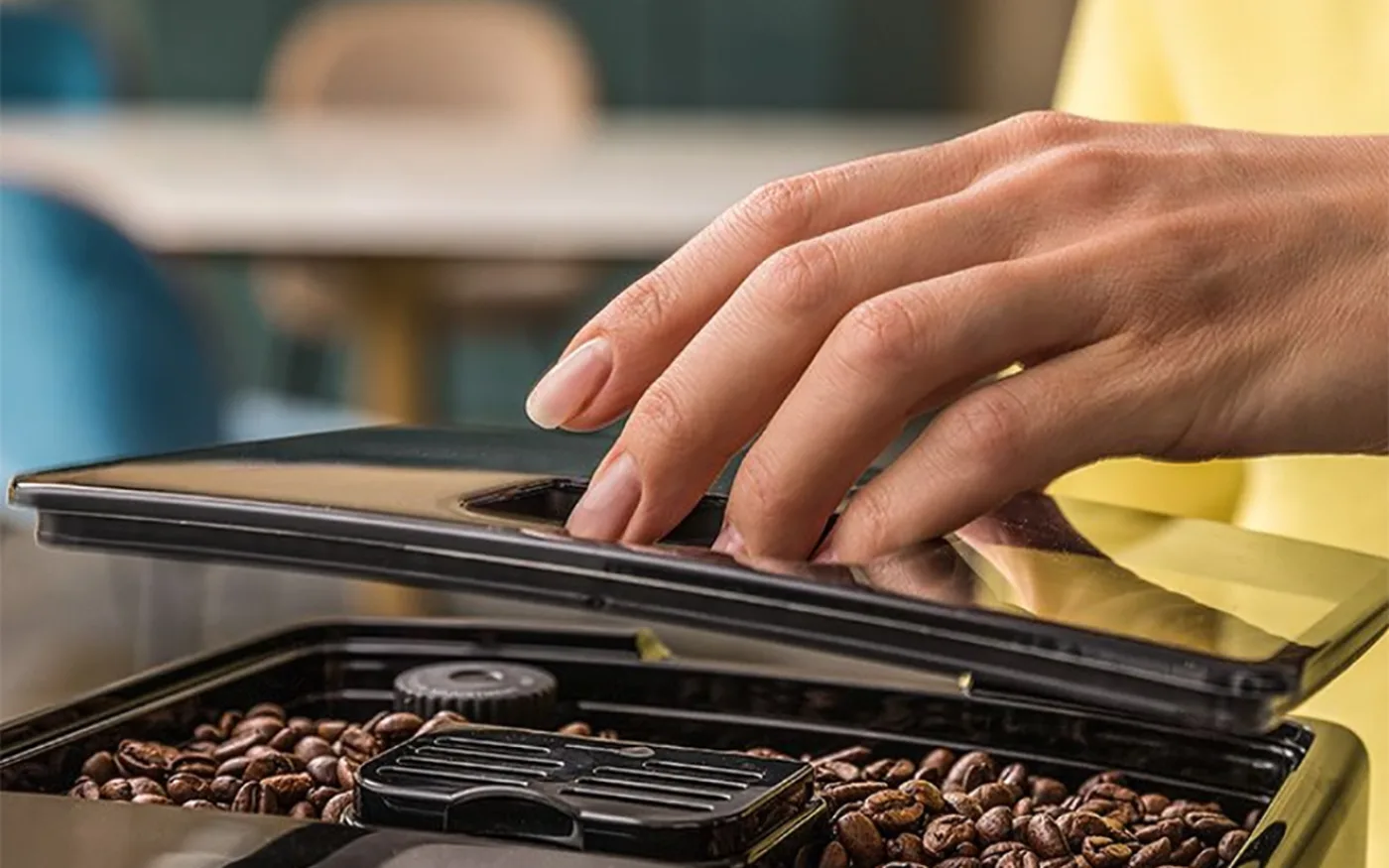 Hand opening the De'Longhi Dinamica bean hopper lid with freshly filled coffee beans