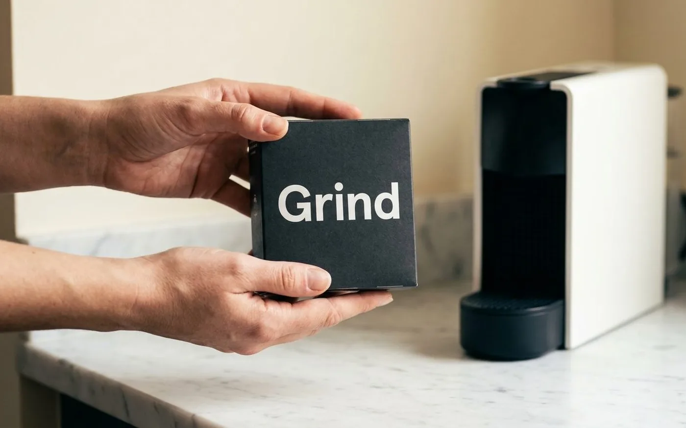Grind coffee pods box held in two hands on white marble counter with Nespresso machine in background