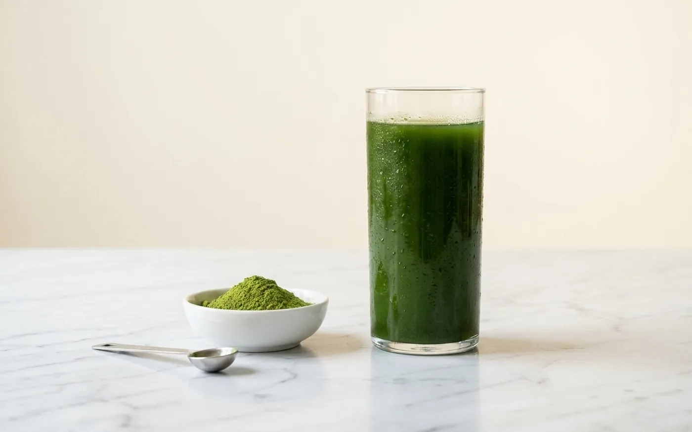 Huel Daily Greens green powder drink in clear glass beside ceramic bowl of emerald green powder on white marble countertop