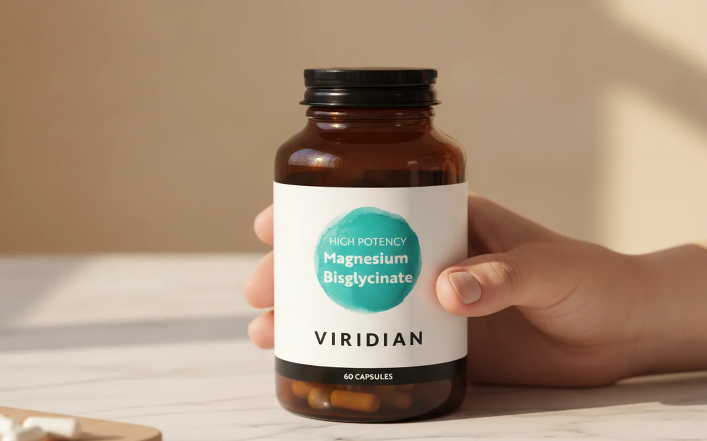 Hand holding an amber bottle of Viridian High Potency Magnesium Bisglycinate capsules on a marble surface