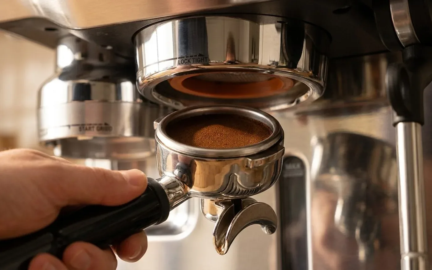 Close-up of the Sage Oracle automatic puck preparation system, a 58mm stainless steel portafilter holding a freshly ground coffee puck below the integrated grinder cradle
